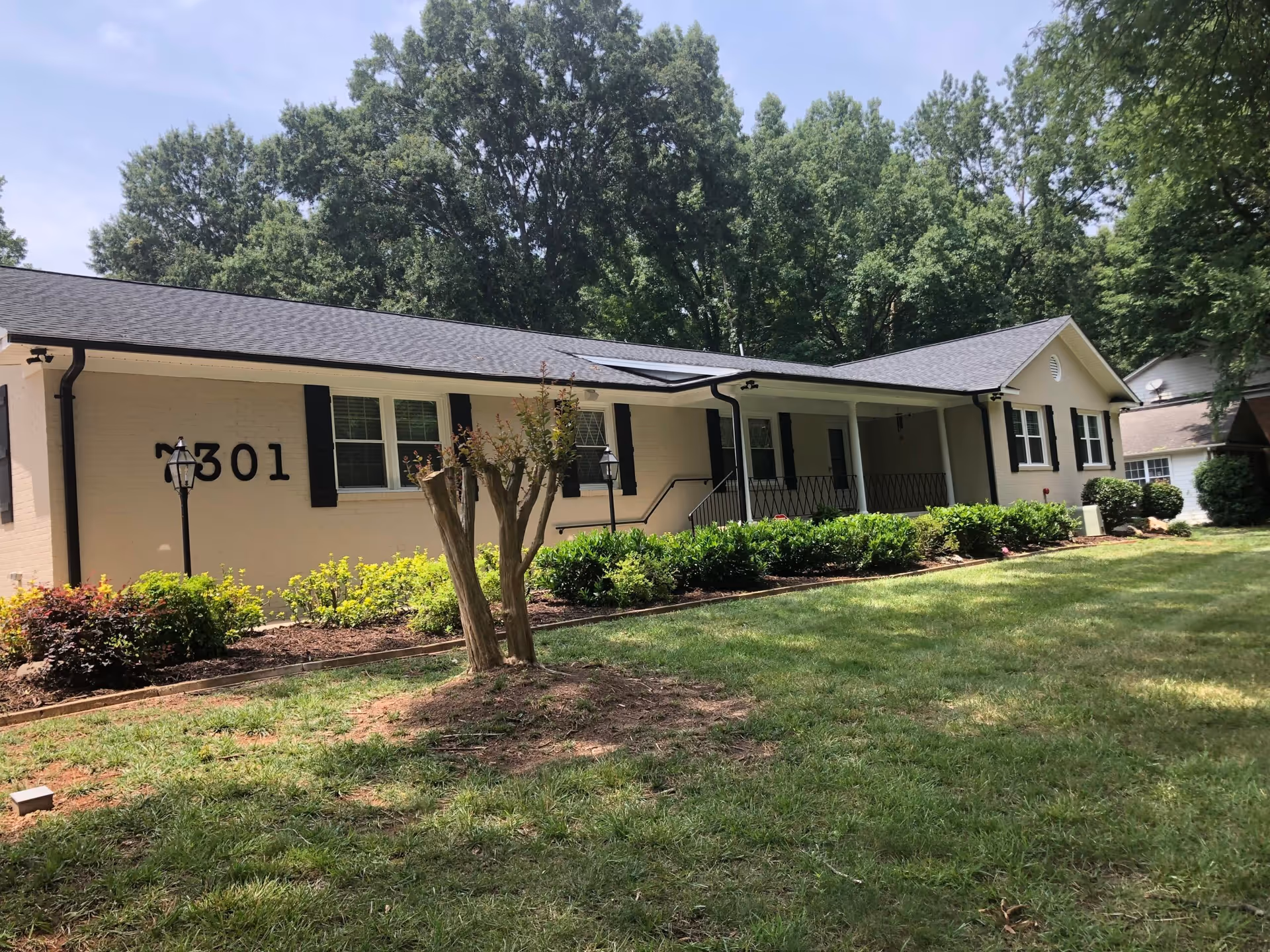 Exterior view of a single-story beige brick building with black shutters and a black roof, surrounded by green bushes and a well-maintained lawn with trees in the background. The building has the number 7301 displayed on the wall.
