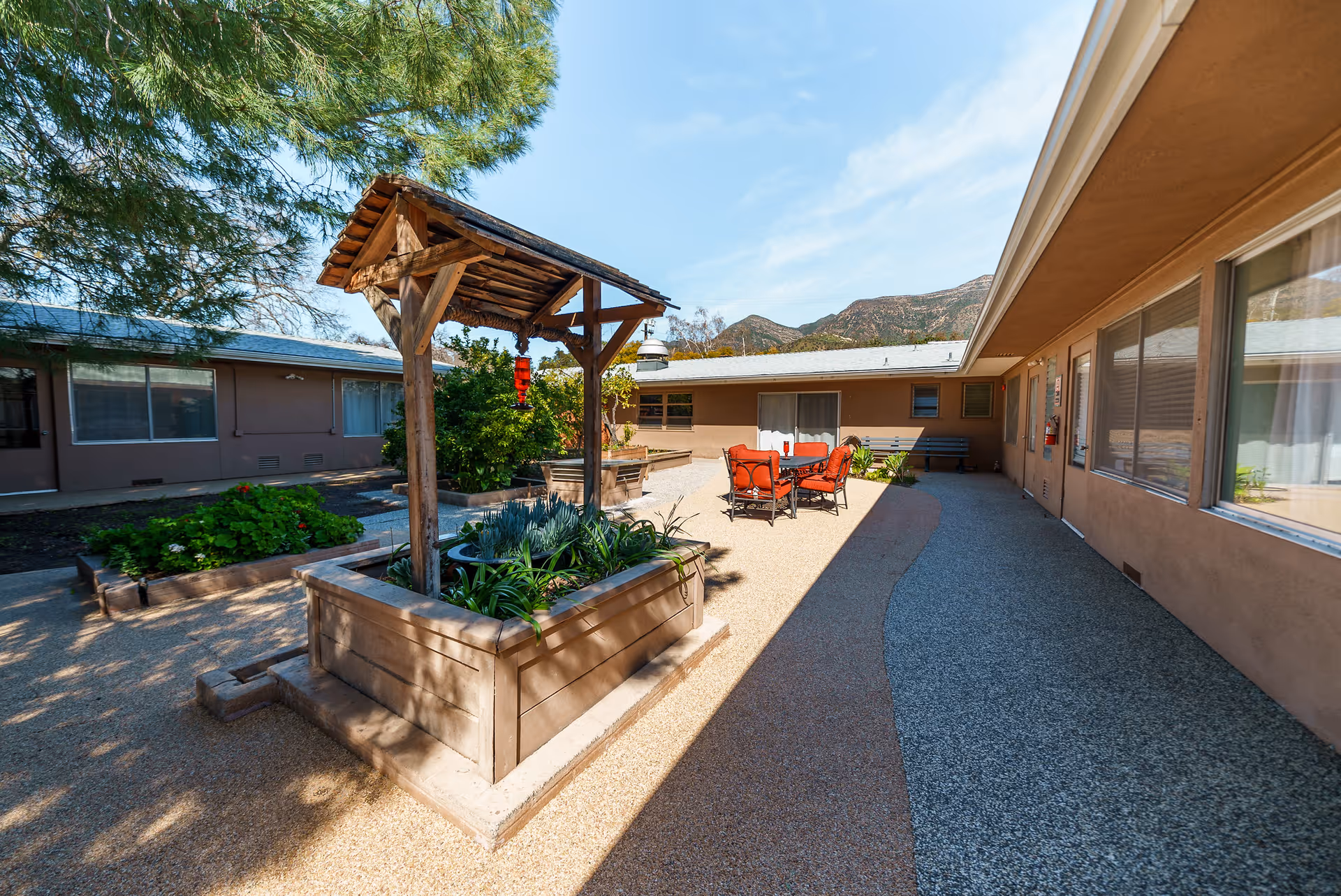 Outdoor courtyard area at Ojai Health & Rehabilitation featuring raised garden beds with plants, a wooden pergola structure, a seating area with red cushioned chairs and a table, surrounded by single-story building walls with windows and doors, and mountains visible in the background under a clear blue sky.
