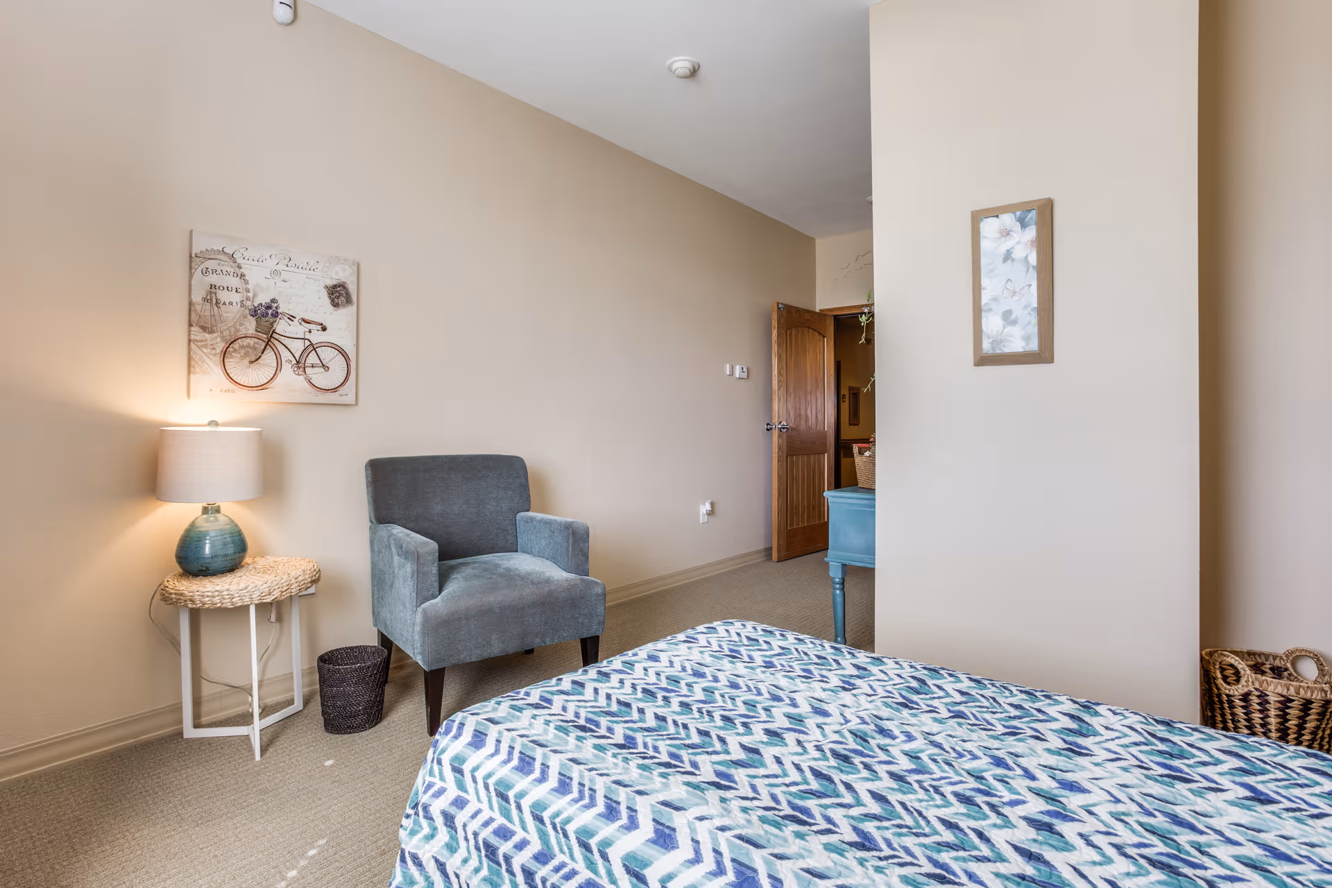 A bedroom with beige walls and carpeted floor featuring a bed with a blue and white patterned bedspread. There is a blue upholstered armchair next to a small round wicker side table with a blue lamp on it. A framed picture of a bicycle hangs on the wall above the chair. A wooden door is open, revealing part of another room with a blue piece of furniture and a wicker basket on the floor.
