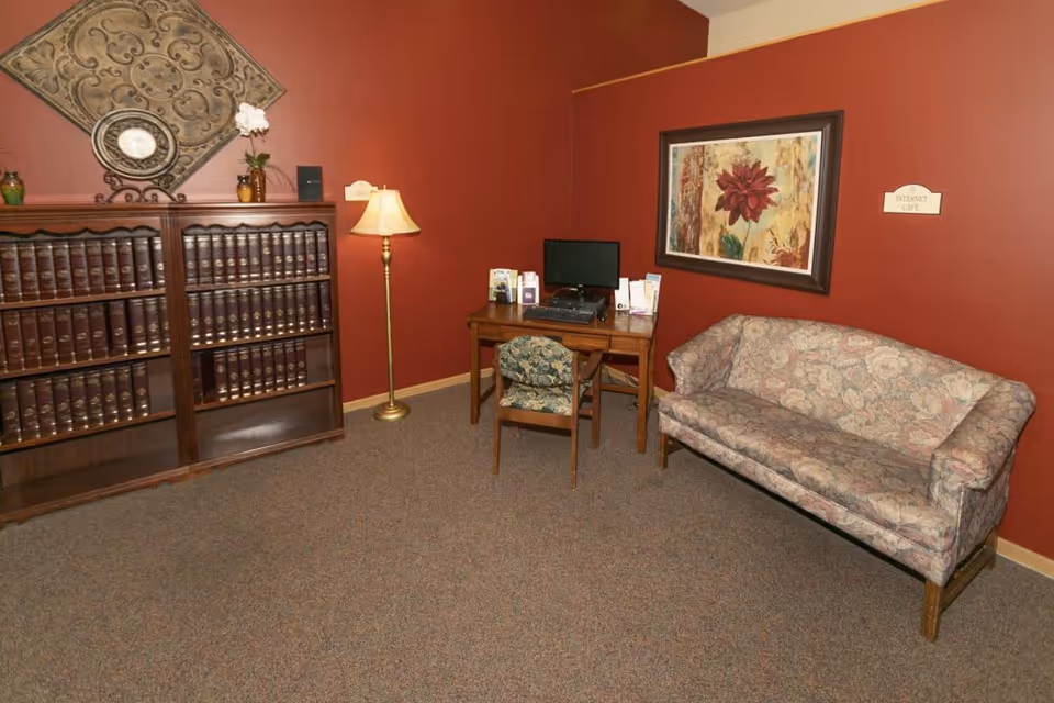 A seating area with a floral sofa, wooden bookshelf, desk with a computer, and a floor lamp against red walls.