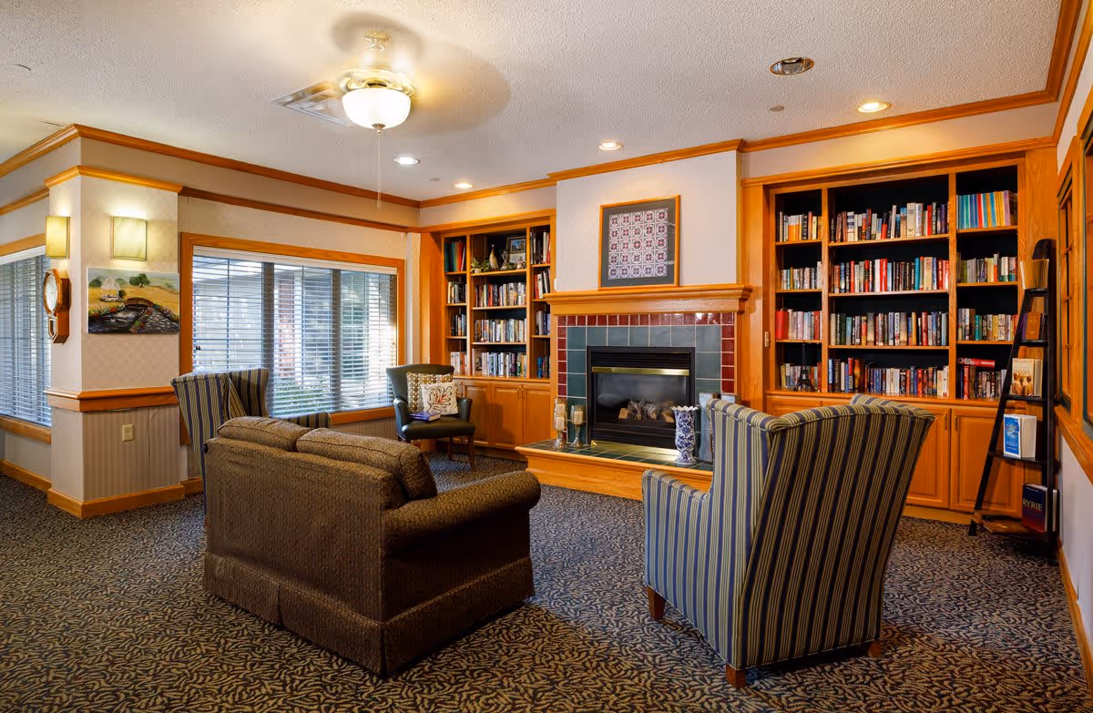 A cozy living room area in an assisted living facility featuring a brown sofa, two striped armchairs, a fireplace with a tiled surround, built-in wooden bookshelves filled with books, a ceiling fan with light, and large windows with blinds allowing natural light to enter.