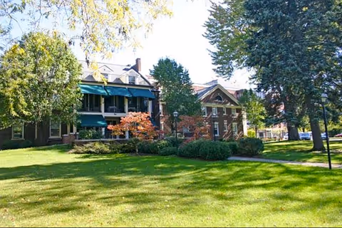 A large brick building with green awnings over the windows, surrounded by trees and a well-maintained grassy lawn under a clear sky.