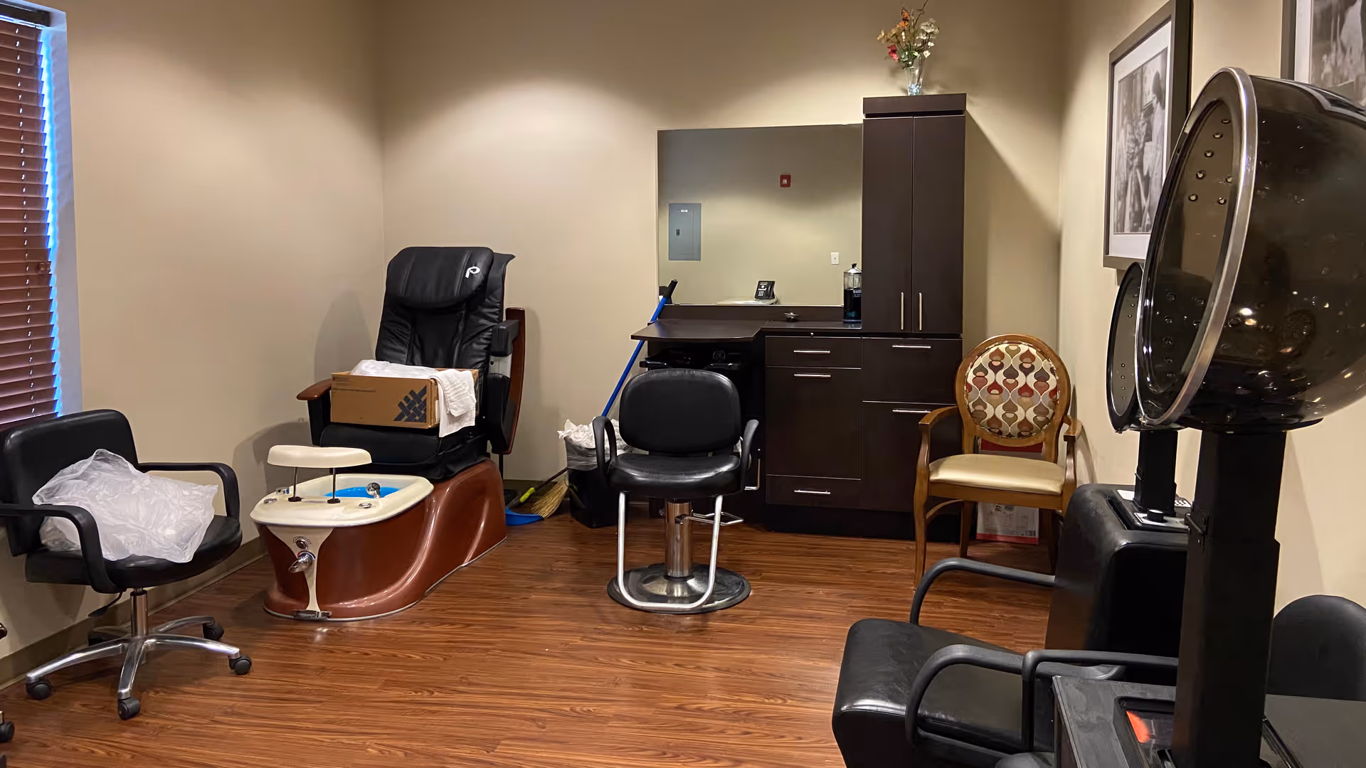 Interior view of a salon or spa room with a pedicure chair, a salon styling chair, a hair dryer chair, and additional seating. The room has wooden flooring, beige walls, a window with blinds, a cabinet with a mirror, and some cleaning supplies in the corner.