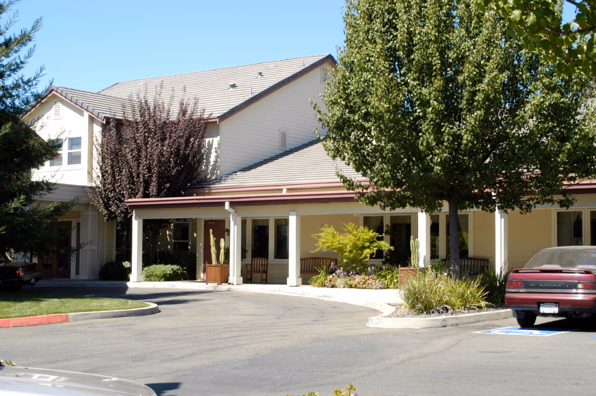 Exterior view of a senior living facility building with a covered porch, several windows, and a few trees and plants in front. There are parked cars and a clear blue sky above.