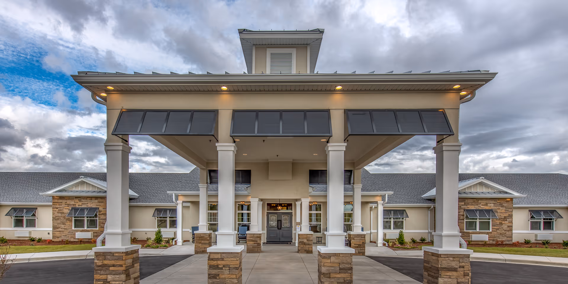 Entrance and front facade of a single-story senior living building with a covered porte-cochere supported by white columns and stone bases under a cloudy sky.