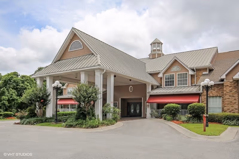 Exterior view of The Waterford at Carpenter's Creek senior living facility showing the main entrance with a covered driveway, red awnings over windows, and well-maintained landscaping including bushes and trees under a cloudy sky.