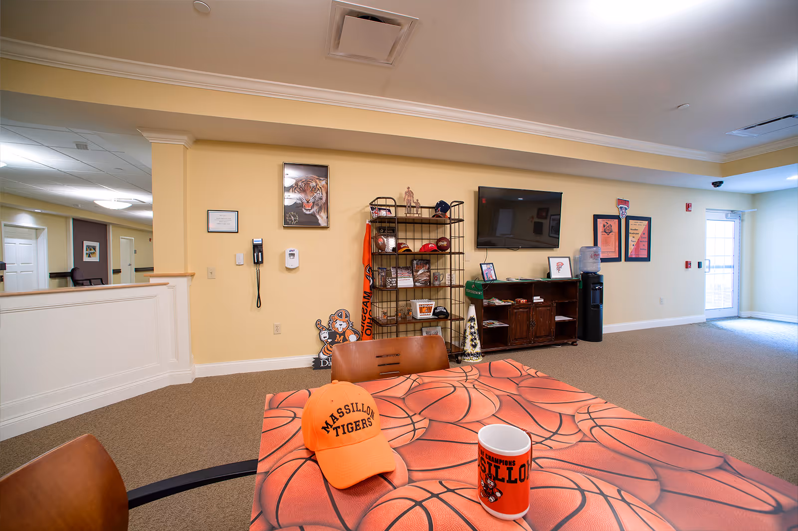 Interior common area of Danbury Massillon facility with a basketball-themed table in the foreground displaying an orange Massillon Tigers cap and a matching mug. The background features a yellow wall with a mounted TV, a metal shelf holding various sports memorabilia, a water cooler, and framed pictures and posters. The room has beige carpet and a door leading outside on the right.