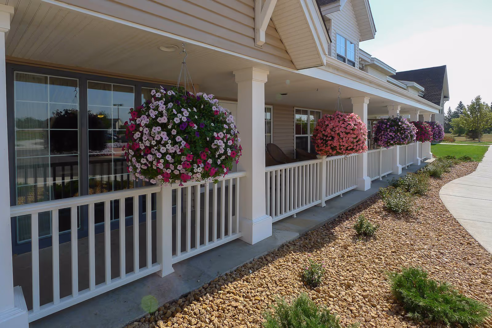 Covered porch area with white railing and hanging baskets of colorful flowers in front of a beige building with large windows. There is a landscaped area with rocks and small plants beside a curved concrete walkway.