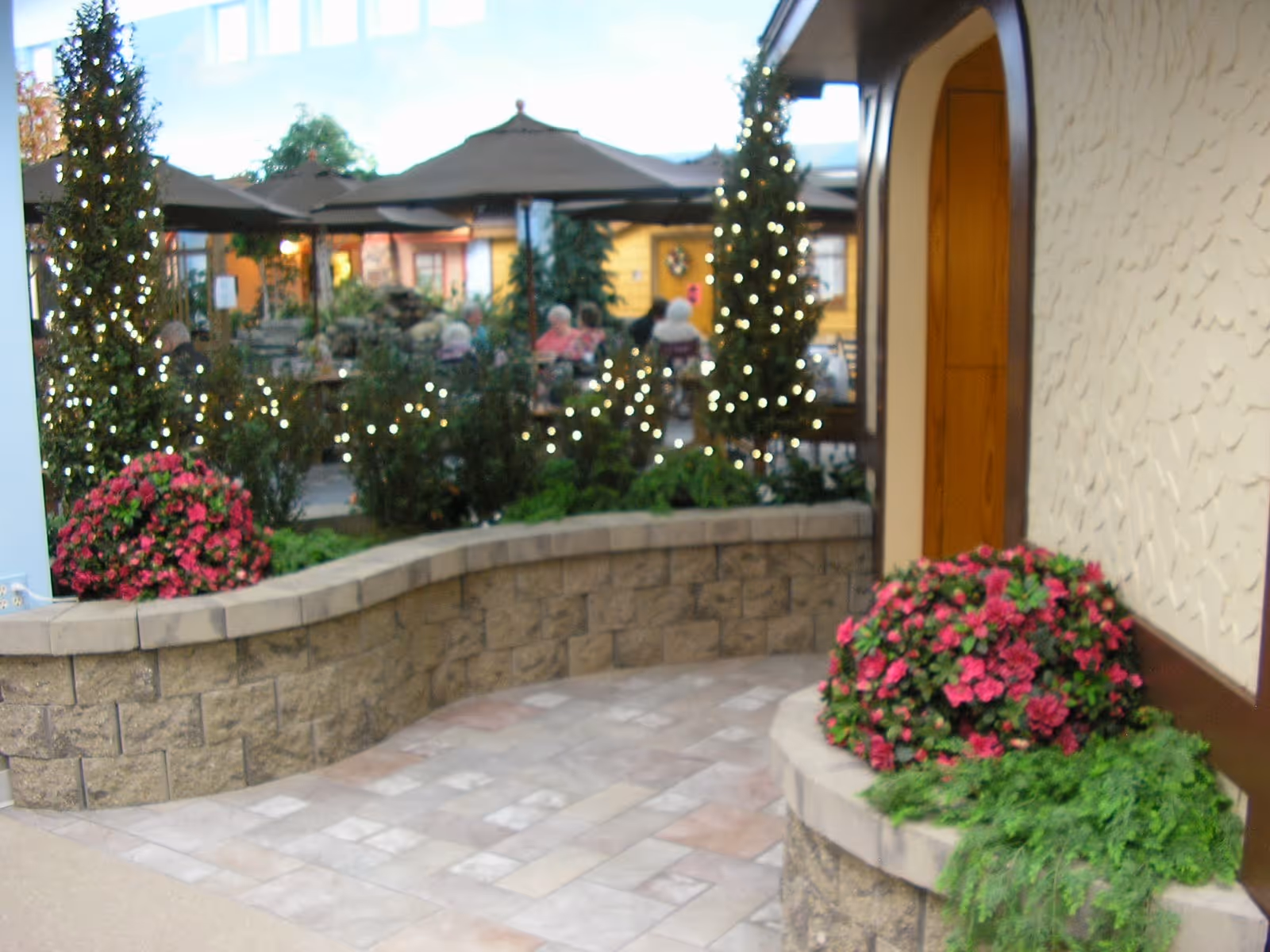 Indoor garden area with stone walls, potted plants with pink flowers, and small trees wrapped in string lights. In the background, there are tables with umbrellas and people sitting, suggesting a communal or dining space inside a facility.