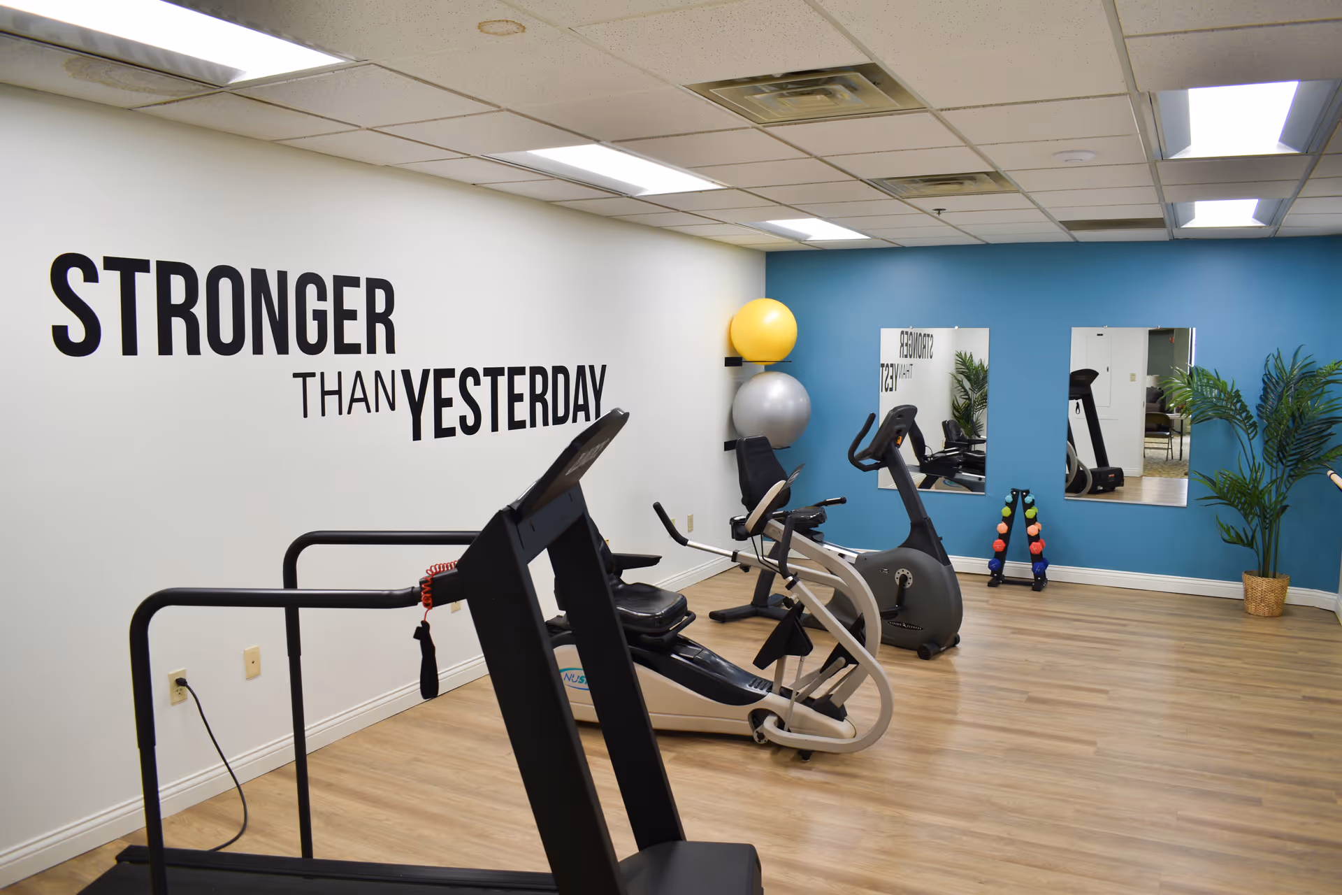 Small exercise room with a treadmill, stationary bikes, exercise balls, dumbbells and a wall decal reading 'STRONGER THAN YESTERDAY'.