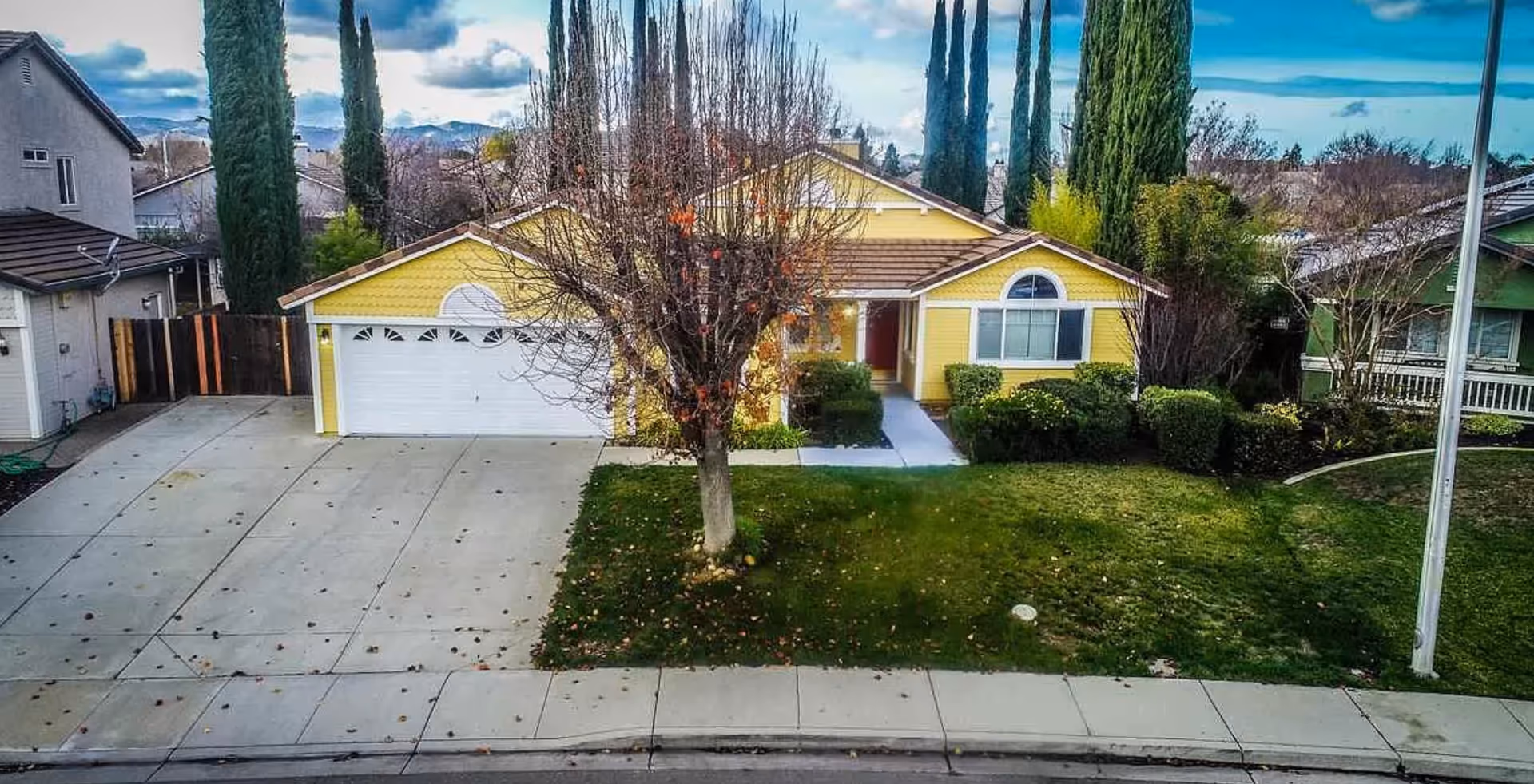 Front exterior view of a single-story yellow house with a two-car garage, a driveway, a leafless tree in the front yard, green bushes, and tall cypress trees in the background under a partly cloudy sky.