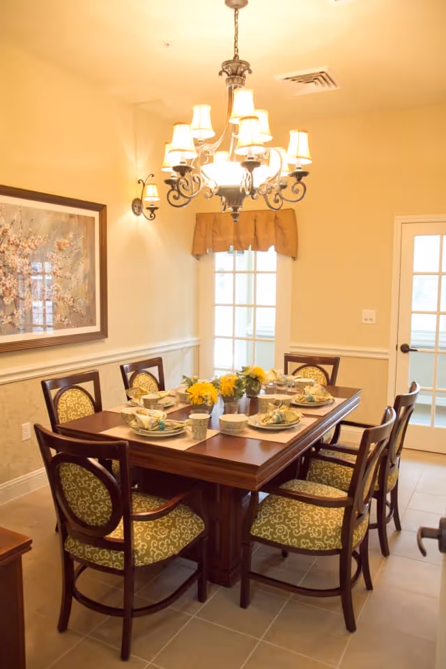 A warmly lit dining room with a wooden rectangular table set for six people. The table has place settings with plates, bowls, cups, and napkins, along with small flower arrangements in the center. The chairs have wooden frames with green patterned upholstery. A chandelier with multiple lampshades hangs above the table, and there is a framed floral painting on the wall. A door with glass panes is visible in the background.