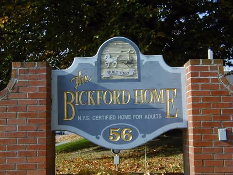 Blue entrance sign reading The Bickford Home between brick pillars at an outdoor entrance.