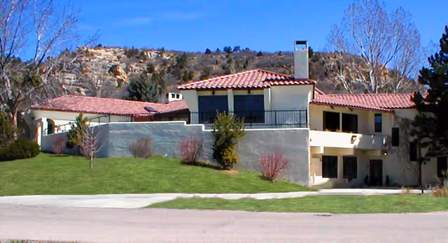 Exterior view of a single-story residential assisted living facility with a red tile roof, beige walls, and a chimney. The building is surrounded by a well-maintained lawn with small trees and shrubs, set against a backdrop of rocky hills and a clear blue sky.