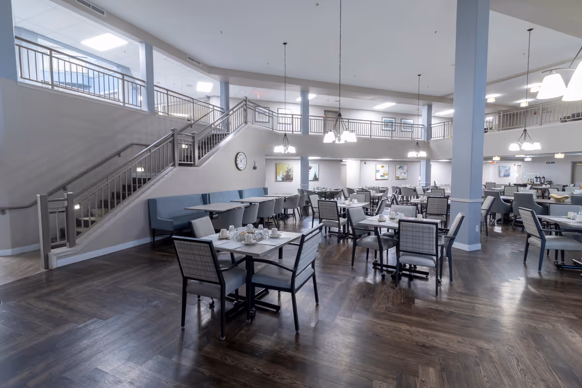 Spacious dining room with multiple tables and chairs arranged neatly. The room features dark wooden flooring, light blue cushioned bench seating along the wall, and several hanging light fixtures. A staircase with metal railings leads to an upper level with additional seating and artwork on the walls.