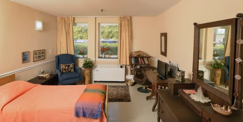Sunlit furnished bedroom with a bed, armchair, desk and dresser with mirror facing windows overlooking greenery.
