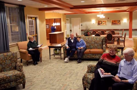 Seniors seated on sofas and chairs in a warmly lit common living room of a continuing care community, with a dining area visible in the background.