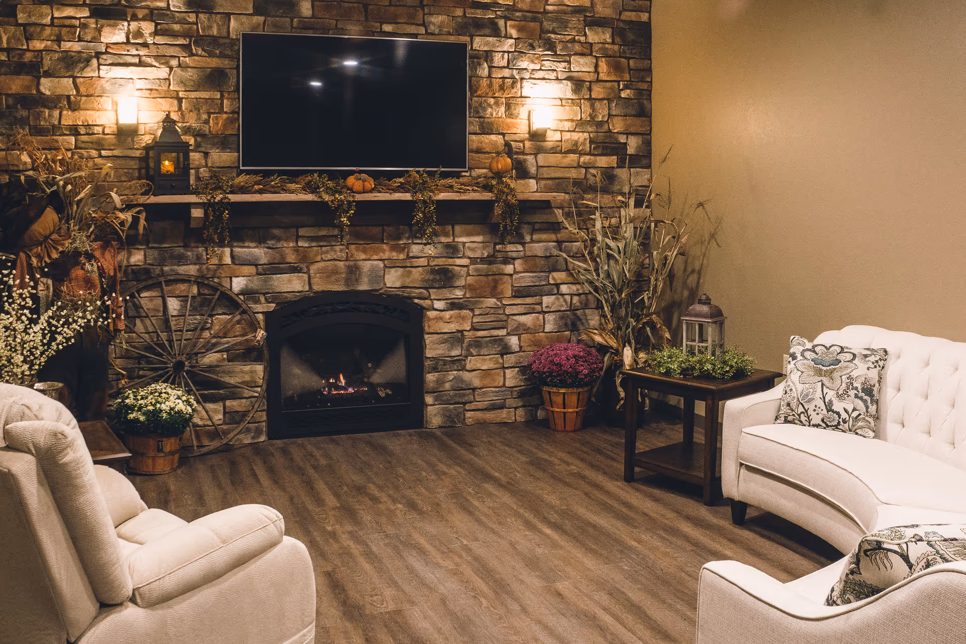 Cozy living room with a stone fireplace, a mounted flat-screen TV above it, and warm lighting. The room features a white armchair and a white sofa with floral cushions, a wooden side table with a lantern and plants, and decorative elements including a wooden wagon wheel, potted flowers, and dried plants.