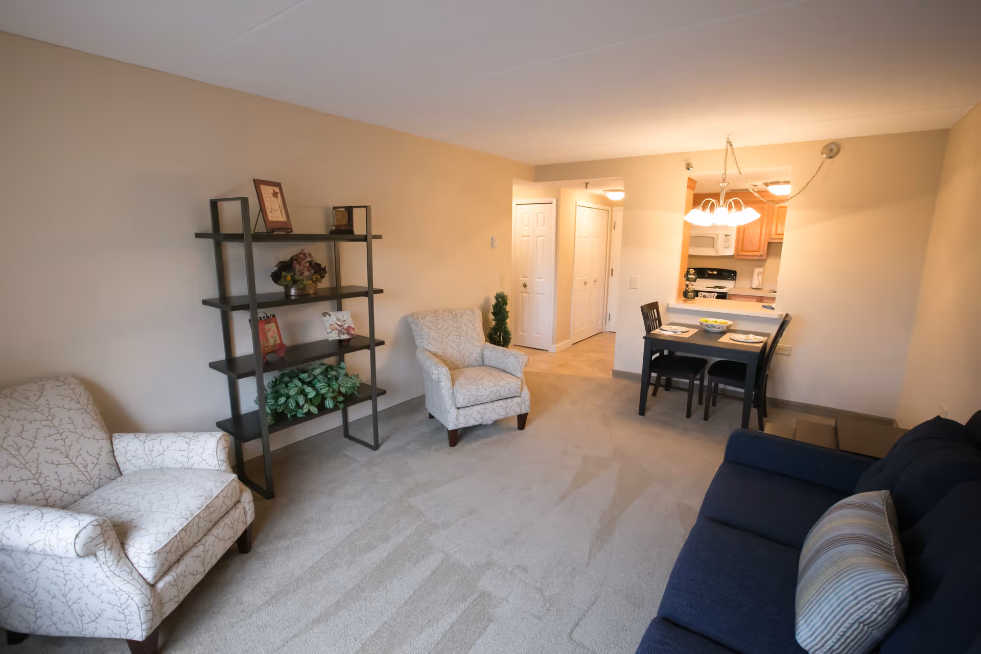 A cozy living room area with two patterned armchairs, a dark blue sofa with a striped pillow, and a black shelving unit holding decorative items and plants. In the background, there is a small dining table set for two with a hanging light fixture above. The kitchen is visible beyond the dining area, featuring wooden cabinets and white appliances.