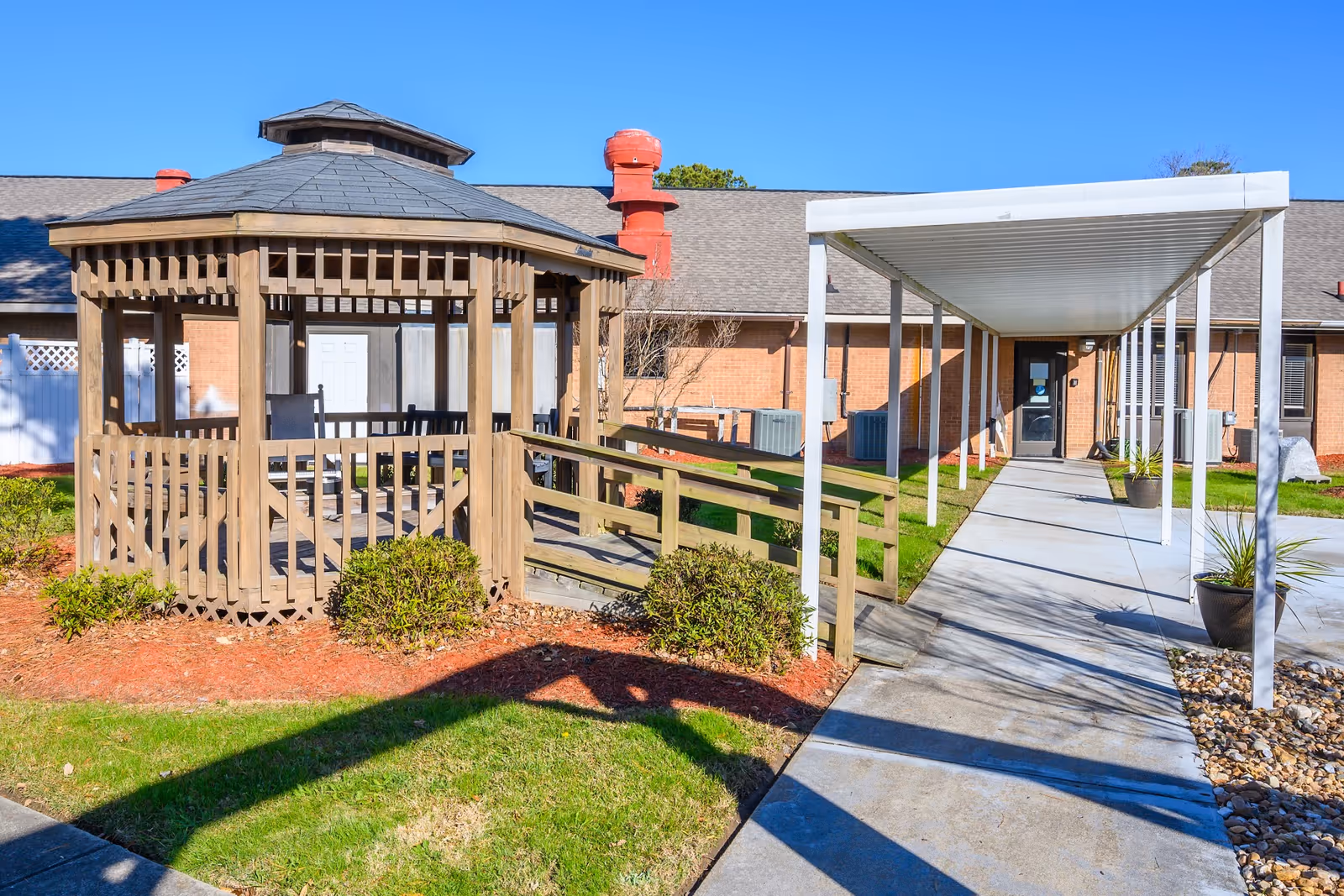 A wooden gazebo and a covered walkway lead to the entrance of a brick rehabilitation center building under a blue sky.