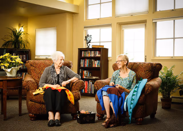Two elderly women sit in upholstered armchairs in a bright common living room, smiling while working on quilts.