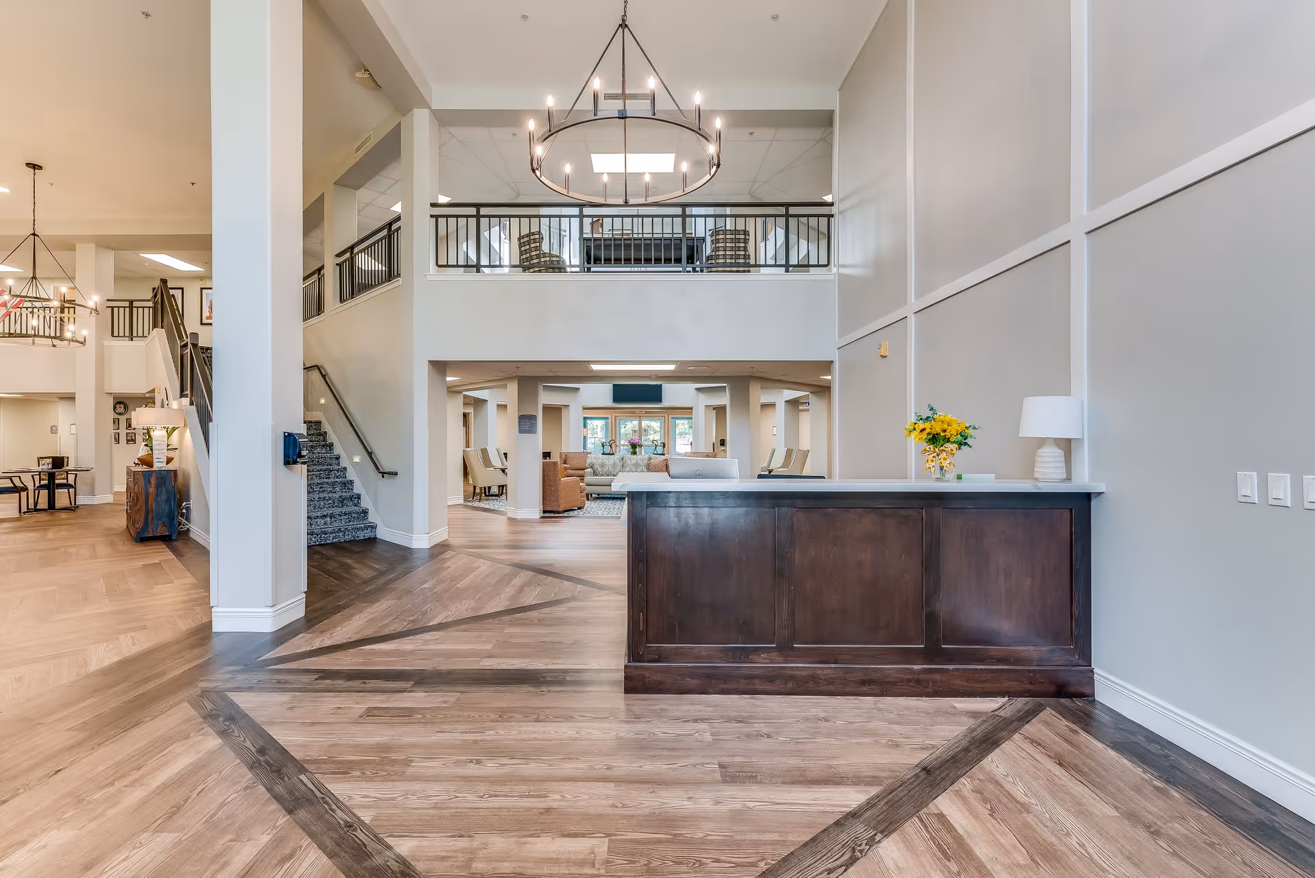 Spacious and well-lit senior living facility interior with a wooden reception desk in the foreground, modern chandelier hanging from the ceiling, staircase to the left, and a comfortable seating area visible in the background.