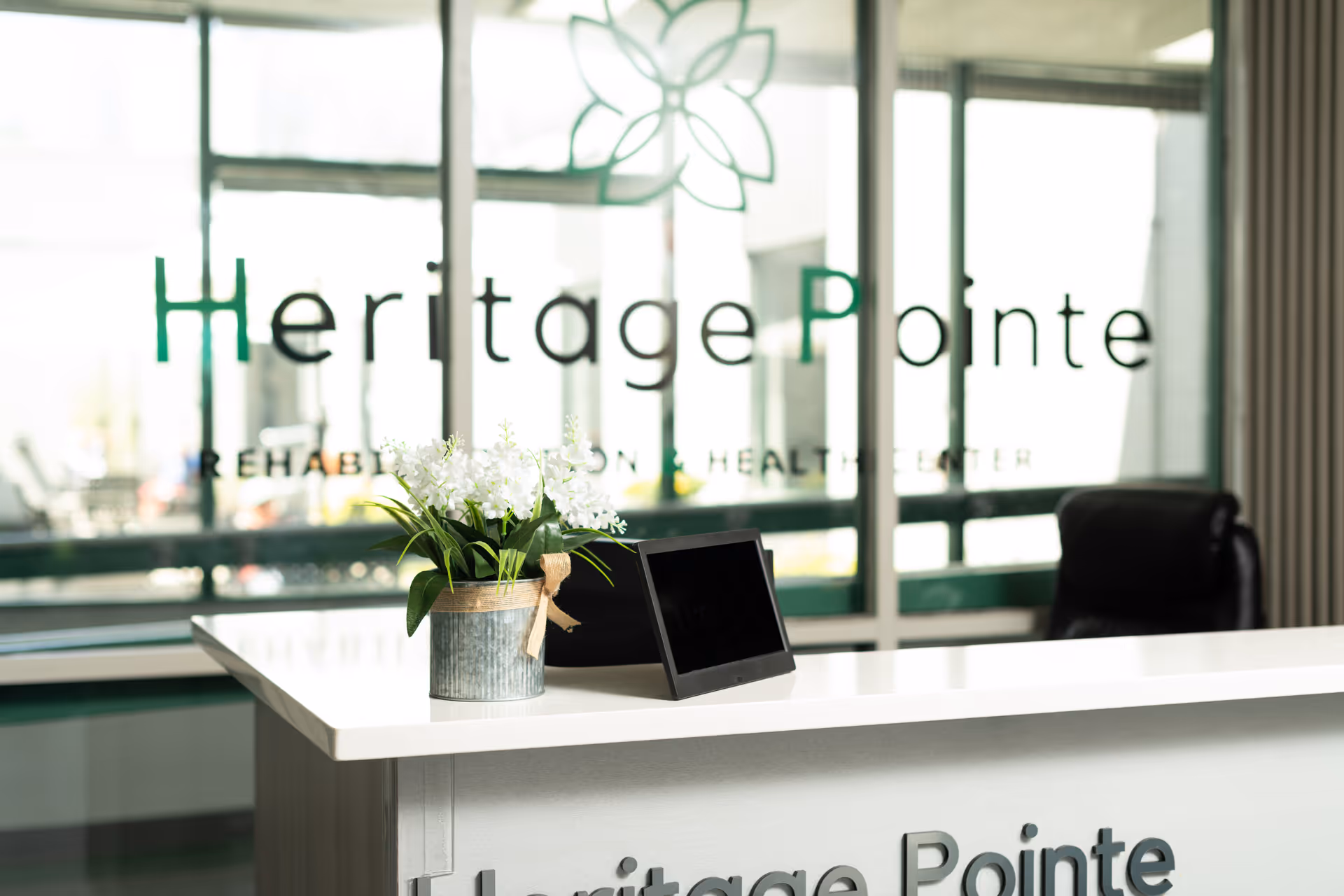 Reception desk with a small potted plant and a digital tablet on it, with a black office chair behind the desk. The glass wall behind the desk displays the text 'Heritage Pointe Rehabilitation and Healthcare Center' with a flower logo above the text.