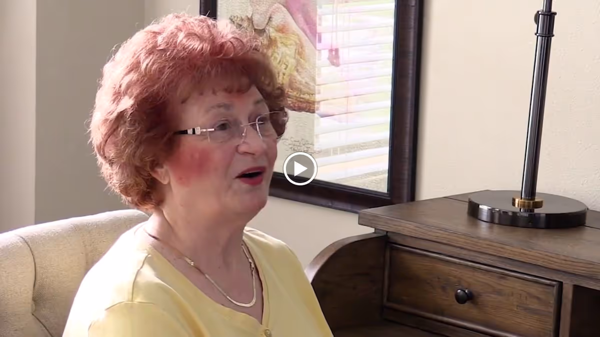 An elderly woman with curly red hair and glasses is sitting on a beige upholstered chair, speaking. She is wearing a yellow top and a gold necklace. Behind her is a wooden side table with a black lamp and a framed artwork on the wall.