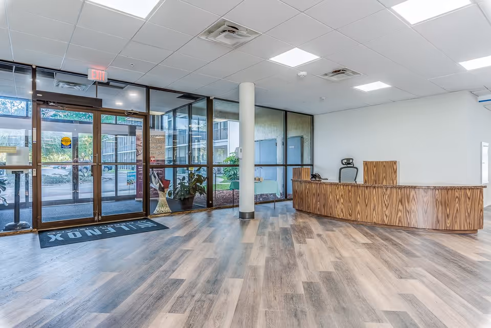 Interior view of a senior living facility lobby with a wooden reception desk, an office chair, large glass entrance doors, and a floor mat with the text 'LENOX'. The floor has a wood-like finish and there are plants visible outside through the glass.