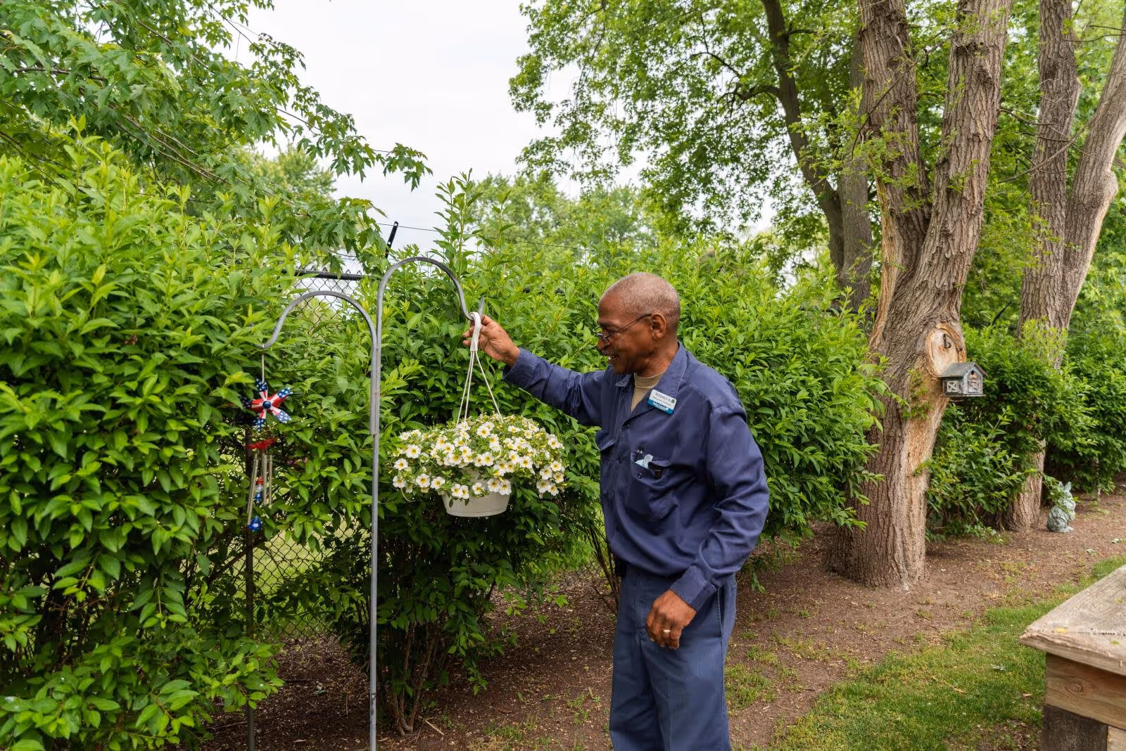 An elderly man wearing a blue uniform is tending to a hanging pot of white flowers in a garden area with green bushes and trees in the background.
