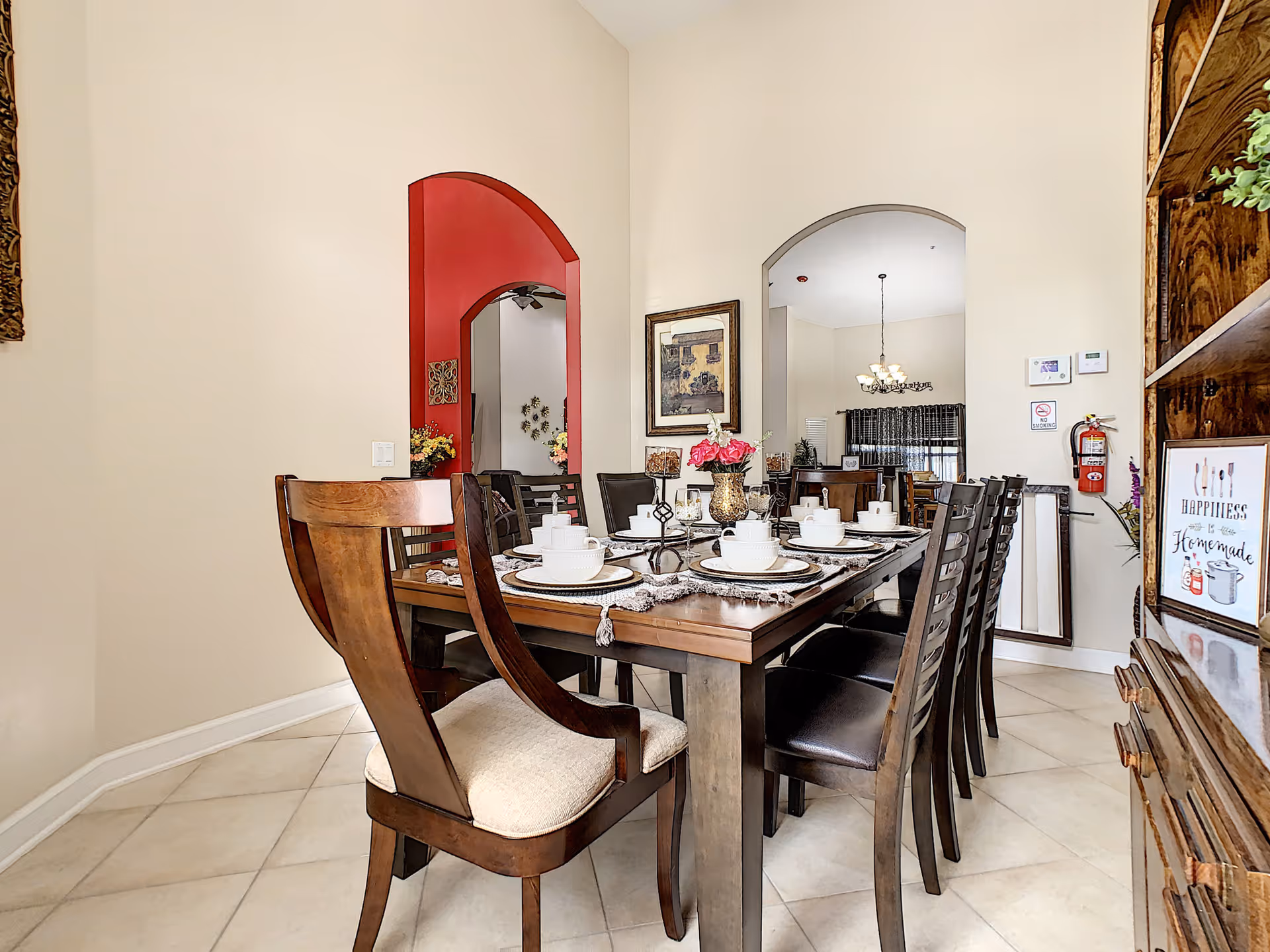 A dining room with a wooden dining table set for eight people with white dishes and cups. The room has beige walls, tiled floors, and two arched doorways, one painted red and the other beige. There is a wooden cabinet on the right with decorative items and a framed sign that reads 'Happiness is Homemade.' A fire extinguisher and thermostat are mounted on the wall near the right archway.