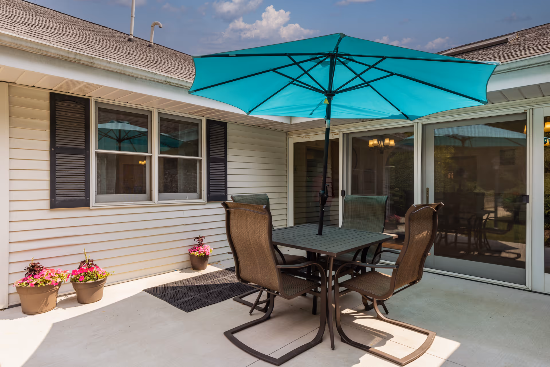 Outdoor patio area with a square table and four chairs under a large turquoise umbrella. The patio is adjacent to a building with white siding, black shutters, and sliding glass doors. There are three flower pots with pink and red flowers placed near the wall.