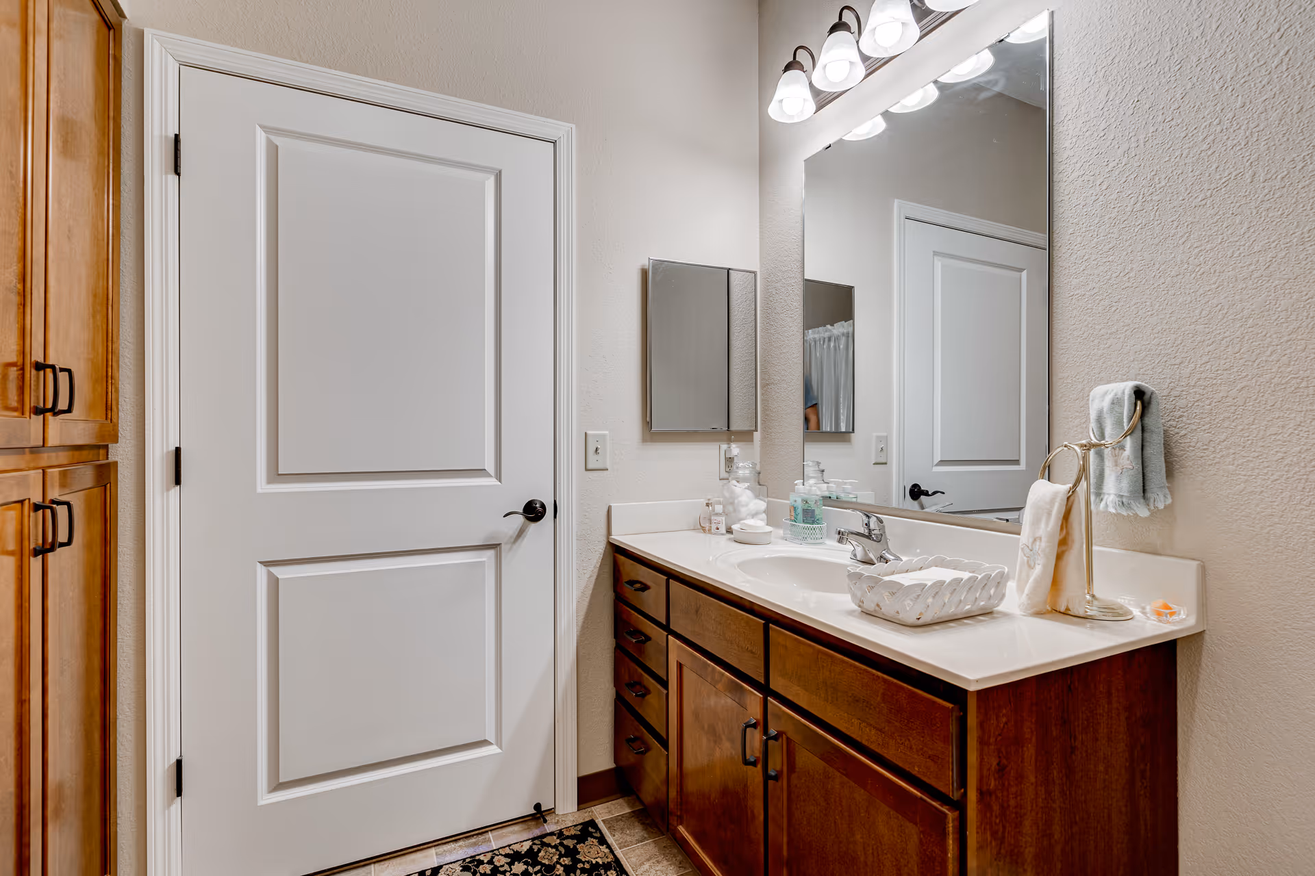 A clean and well-lit bathroom featuring a white door, wooden cabinets with dark handles, a countertop with a sink, soap dispenser, cotton balls, and a towel on a stand. Above the sink is a large mirror with three light fixtures.