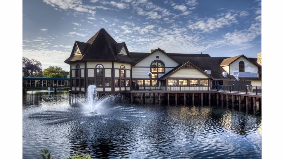 A large building with a steep roof and multiple windows sits on stilts above a body of water with a fountain spraying water in the center. The sky is partly cloudy with soft sunlight illuminating the scene.
