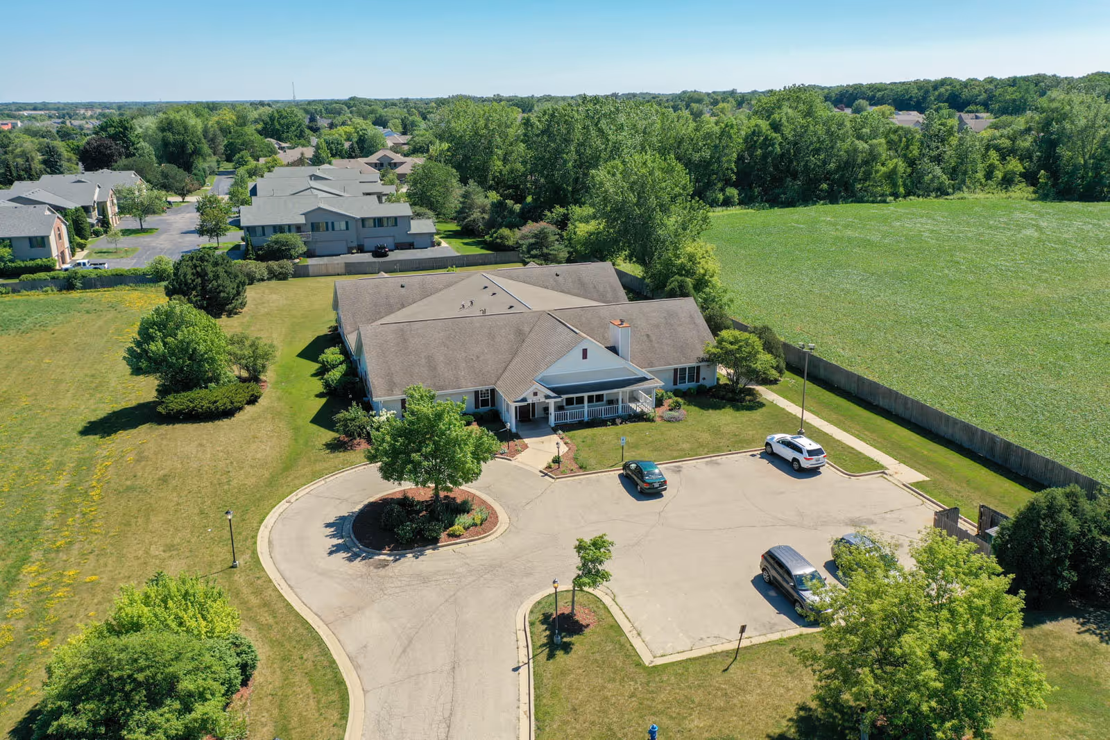 Aerial view of a single-story senior living building with a circular driveway, parked cars, surrounding lawns, trees, and nearby houses and fields.