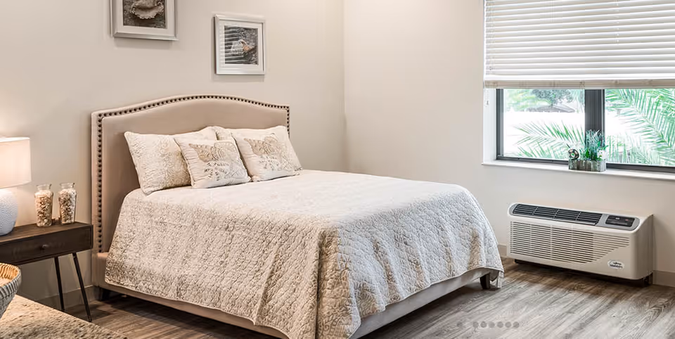 Bright neutral bedroom with a neatly made bed, nightstand with lamp, and a window air-conditioning unit.