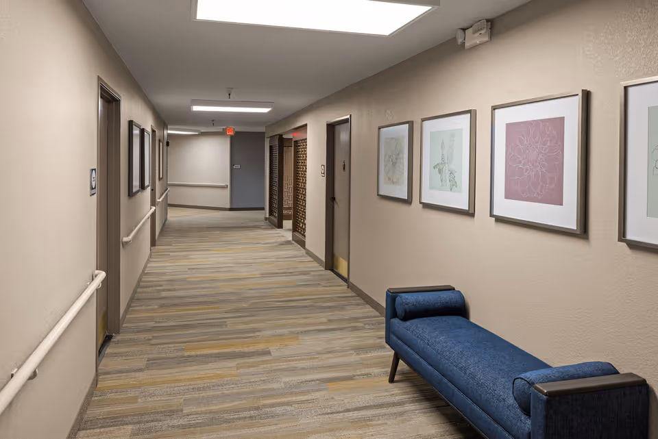 A well-lit hallway in a senior living facility with beige walls, handrails along both sides, framed botanical artwork on the right wall, and a blue cushioned bench. Several closed doors line the hallway, and the floor is covered with a patterned carpet in neutral tones.