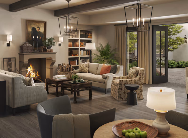 Cozy living room with sofas and armchairs arranged around a coffee table and lit fireplace, bookshelves, lamps, and doors opening to an outdoor patio.