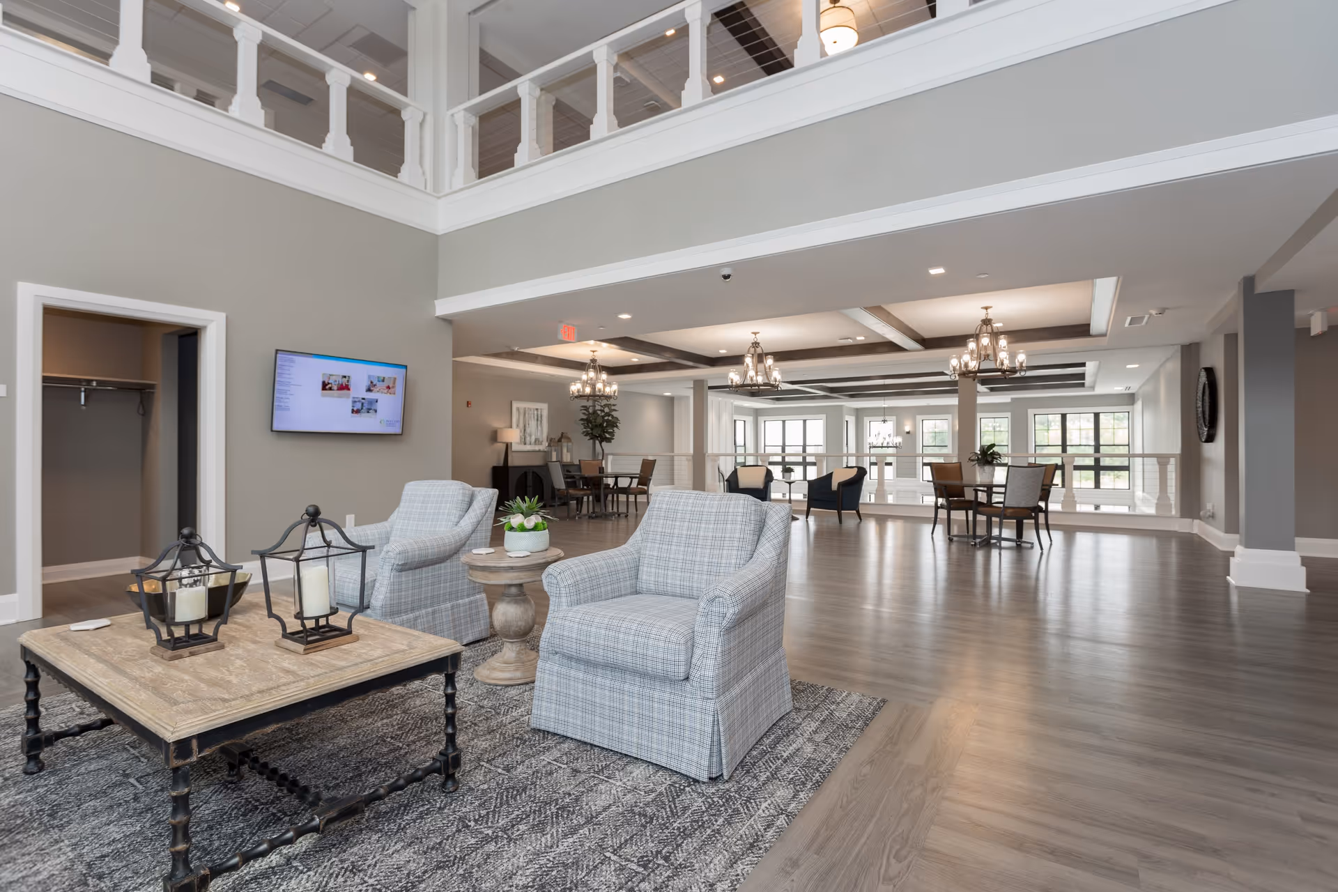 Spacious senior living facility common area with high ceilings and a mezzanine level. The room features two plaid armchairs and a wooden coffee table with decorative lanterns and a small plant. In the background, there are dining tables and chairs under elegant chandeliers, large windows letting in natural light, and a wall-mounted TV.