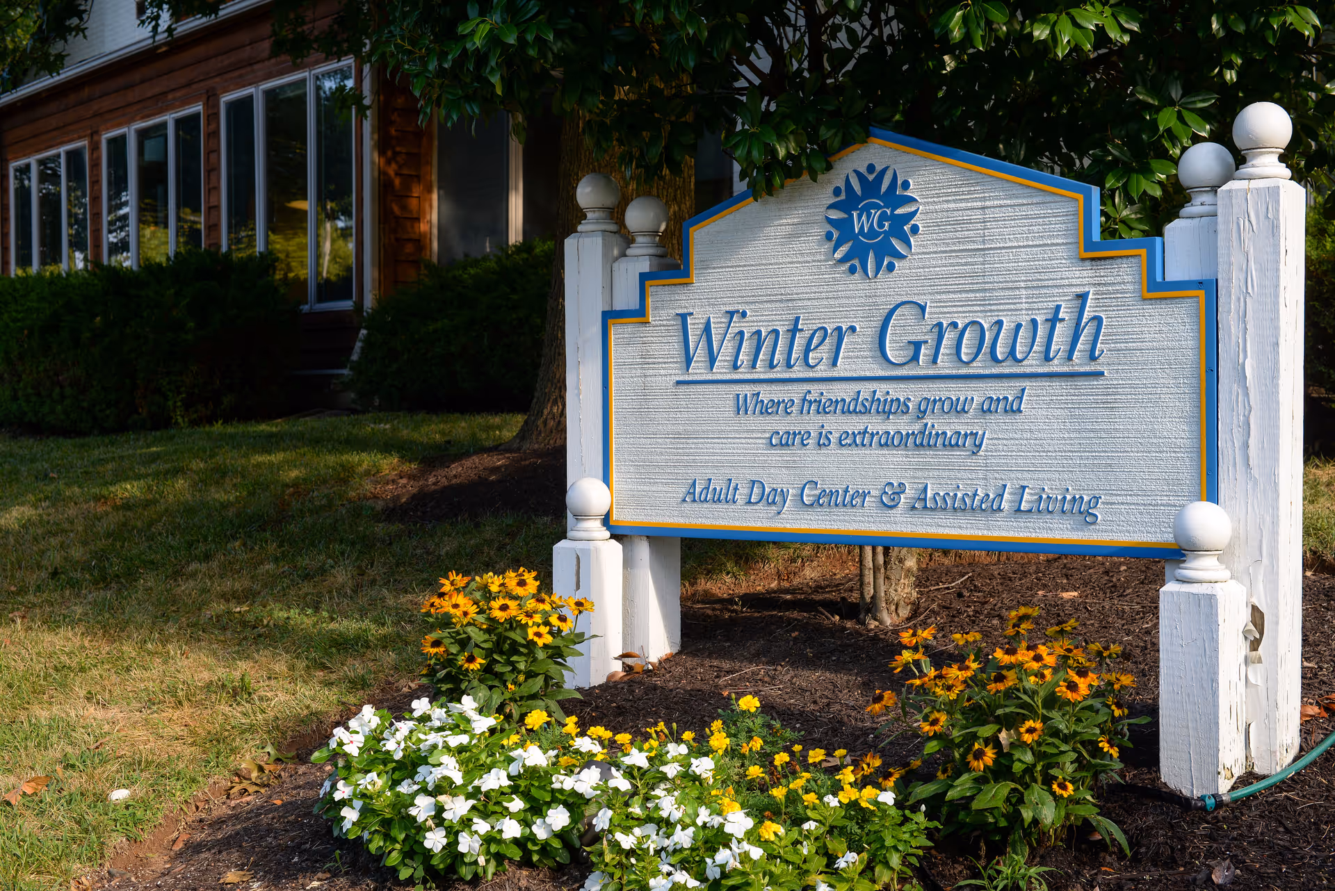 Outdoor entrance sign reading "Winter Growth" for an adult day center and assisted living, set in a landscaped bed with flowers and a building in the background.