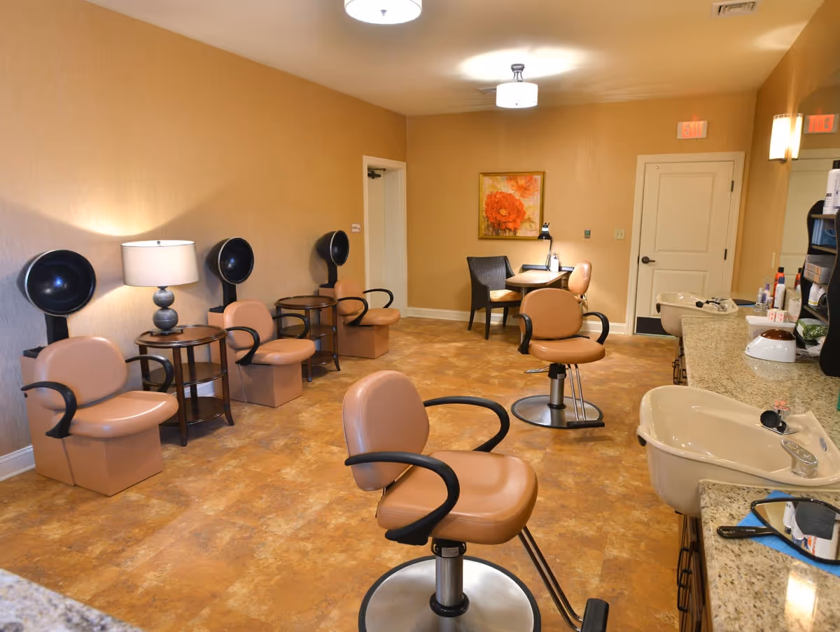 Interior view of a salon area in an assisted living facility with several tan salon chairs, hair dryers, a table with chairs, sinks, and a countertop with hair care products. The walls are beige with a floral painting and there are ceiling lights illuminating the room.