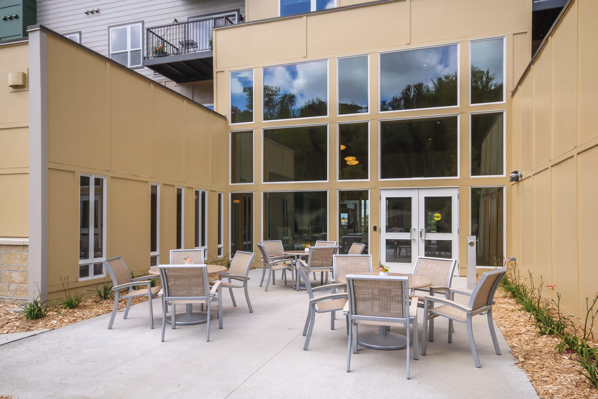 Outdoor patio area at Woodland Hill facility with several round tables and chairs arranged on a concrete surface, surrounded by beige building walls with large windows reflecting the sky and trees.