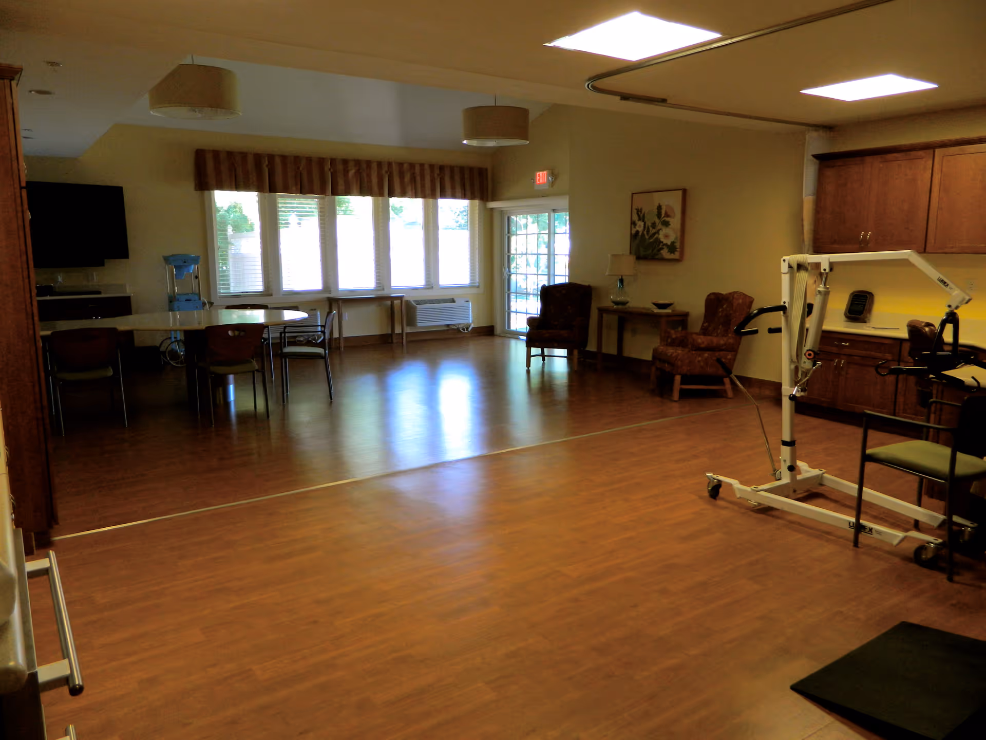 A spacious room with wooden flooring and beige walls, featuring a table with chairs near large windows with blinds, two upholstered armchairs with a small table and lamp between them, a wall-mounted TV, a water dispenser, and a mechanical patient lift device near wooden cabinets.