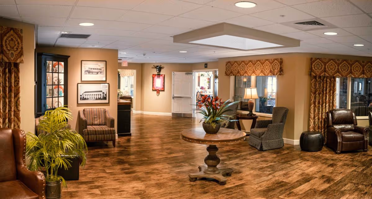 A spacious and warmly decorated common area in a senior living facility featuring wood flooring, multiple comfortable armchairs, a round wooden table with a potted plant centerpiece, framed pictures on the walls, large windows with patterned valances and curtains, and soft lighting from table lamps.