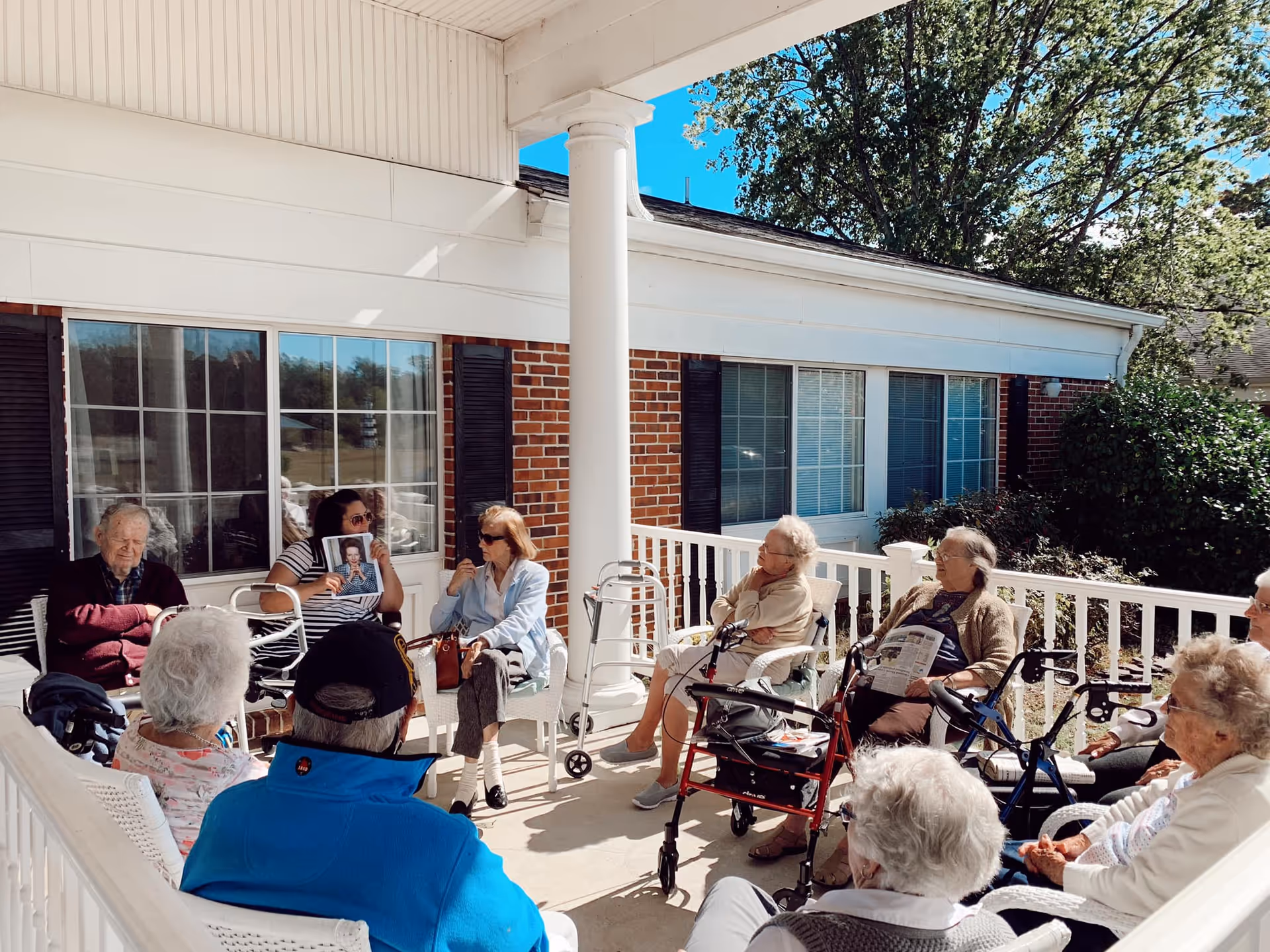 A group of elderly people sitting in a circle on a covered porch outside a brick building, some using walkers, engaging in conversation on a sunny day.