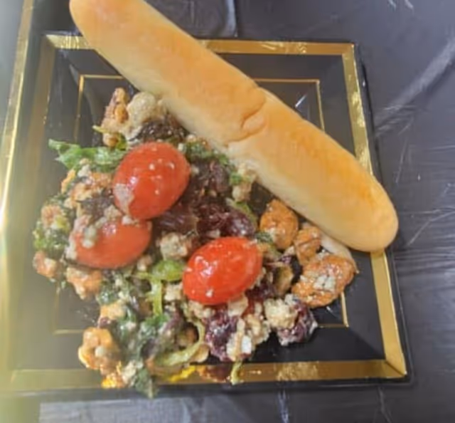 A black square plate with a gold border containing a salad made of leafy greens, cherry tomatoes, walnuts, and crumbled cheese, accompanied by a breadstick on the side.