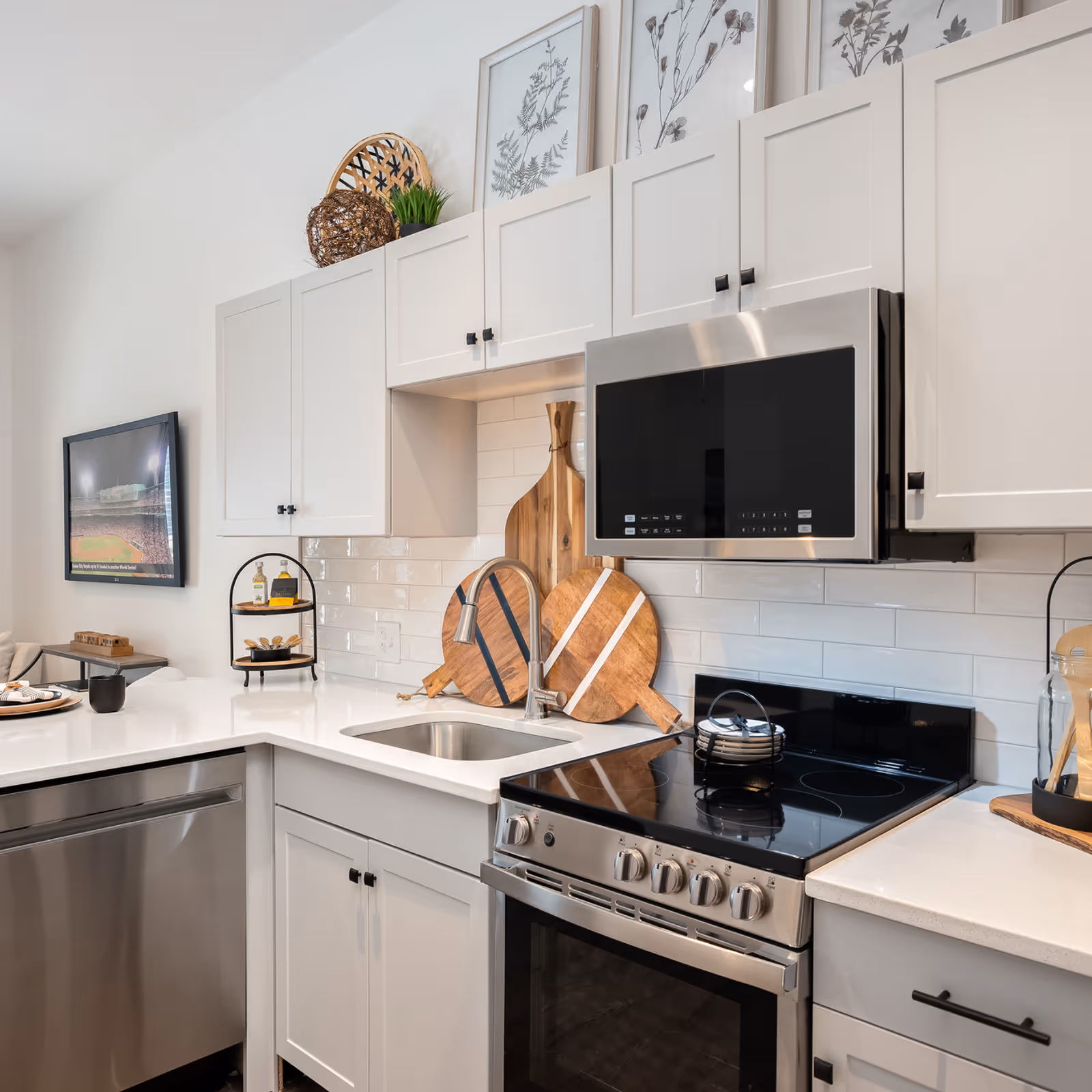 Modern kitchen with white cabinets, stainless steel appliances including a microwave and stove, a small sink, and decorative wooden cutting boards and framed botanical prints on the wall.