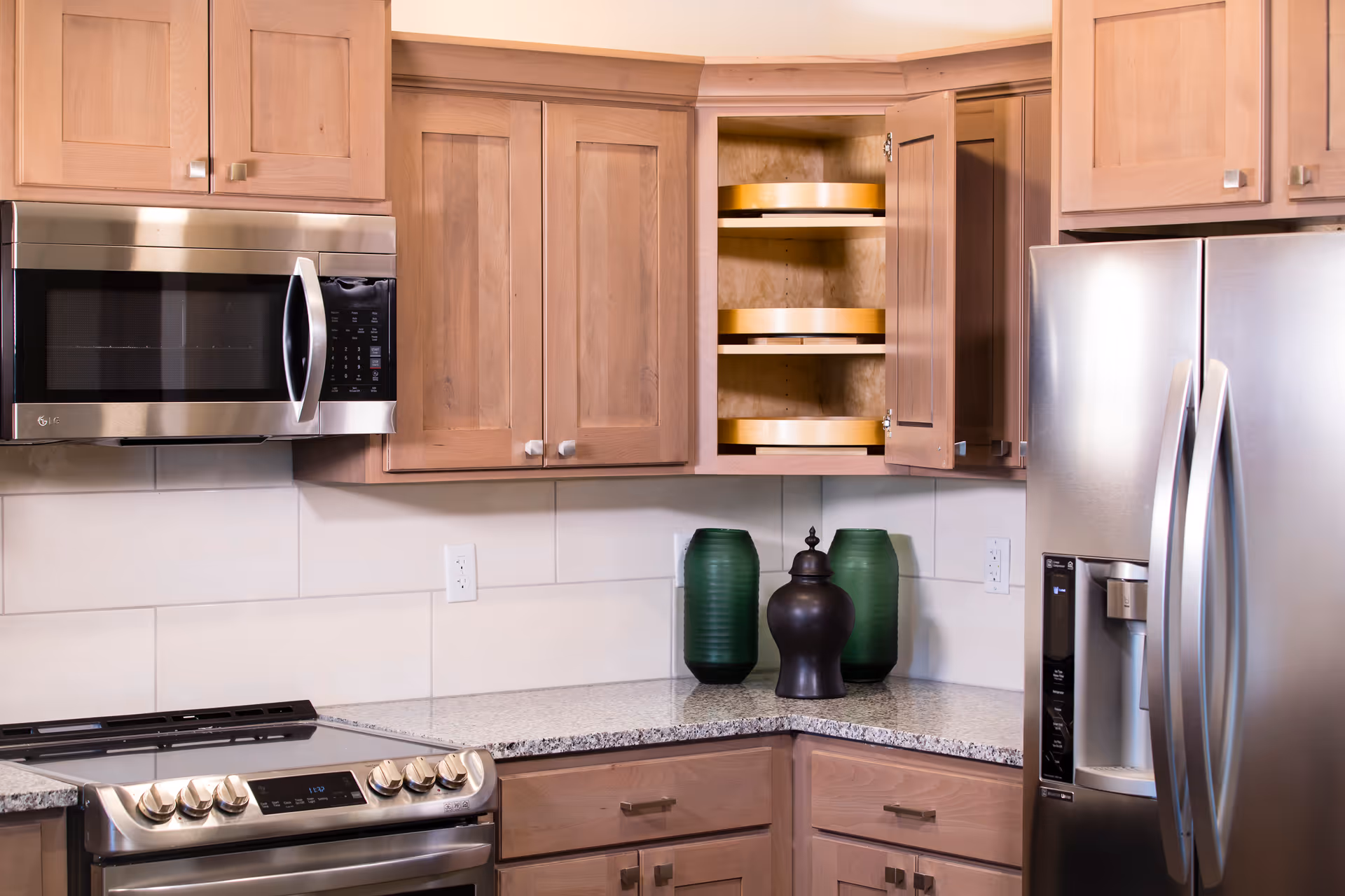 Modern kitchen with light wood cabinets, a stainless steel microwave, stove, and refrigerator. The corner cabinet door is open, revealing rotating shelves. Two green vases and one black decorative jar are placed on the granite countertop.