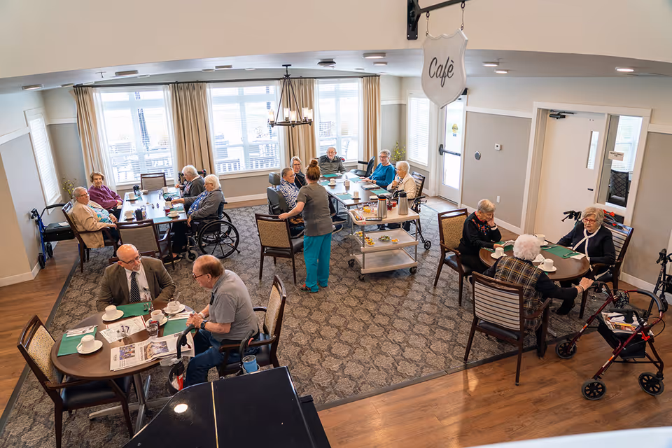 A dining area in a senior living facility with several elderly residents seated at round and rectangular tables, some using walkers and wheelchairs. A staff member is serving or assisting at one of the tables. The room has large windows with beige curtains, a chandelier, and a sign hanging from the ceiling that reads 'Cafe'.