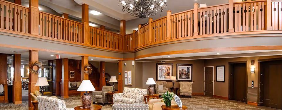 Spacious two-story senior living lobby with wooden balcony railings, a seating area of sofas and armchairs, lamps, and decorative accents.